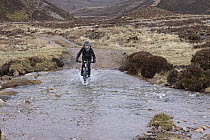 Boy on mountain bike cycling through water, north of Loch Eanaich, Cairngorms, Scotland, UK. April, 2023. Model released.