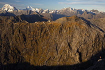 A biodiversity ranger standing in the last rays of sunlight on an alpine ridge of Mount Danger with mountains in background, near Milford Sound, Fiordland National Park, South Island, New Zealand. May...