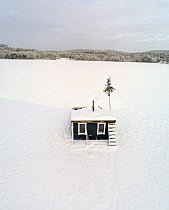 Small cottage at edge of frozen lake covered in snow with forest in background, Odalen, Innlandet, Norway. January, 2018.