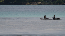 Young fishermen using traditional wooden canoe and net, in shallow water, to catch fish. A child is walking through the water, playing with stones. Near Kavieng, New Ireland, Papua New Guinea.