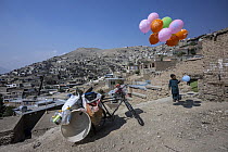 Boy holding bunch of balloons standing near his bike laden with items he is selling, Hope Cabul, Afghanistan. October, 2023.