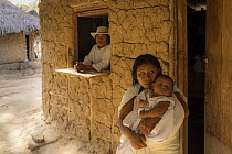 Woman holding her baby with man looking out of window behind in village in the El Encanto Wiwa community, Sierra Nevada de Santa Marta, Colombia. December, 2021.