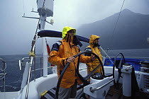 Two men in wet-weather gear helming 88ft sloop "Shaman" as she motors through Fjordland, South Island, New Zealand 2001