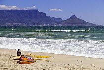 A windsurfer taking a break on a sandy beach overlooking Cape town, Table Mountain and Lion's Head, South Africa.