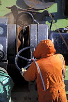 A member of the crew on a commercial ship letting down the anchor using the ship's windlass.