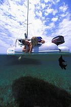 Split-level view of two children looking over the edge of a tender into the water with a catarmaran anchored behind, Belize. Model released.