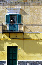 Old woman watching from balcony window overlooking main square, Ventotene Island, Pontine Islands, Italy.