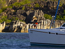 Woman sat reading a book on the bow of a cruising catamaran anchored by eroded granite boulders that line the shores of the Seychelles, Indian Ocean.