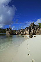 Naturally carved granite boulders line the shore of a white sandy beach with footprints in the sand, Seychelles, Indian Ocean.