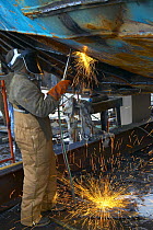 Worker in a shipyard working on the hull of a trawler with an acetelene torch.