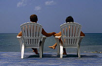 Couple sitting on plastic chairs on a sun deck, holding hands and looking out to sea.