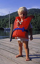 A little boy standing on a jetty wearing a life jacket.
