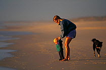 Mother helping young child to walk towards the sea with a dog behind them. Model released.