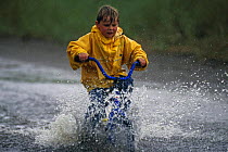 Young boy in a yellow waterproof coat, cycling through a big puddle of water. Model released.