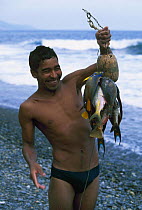A fisherman in swimming trunks holding up his catch of fish on a pebble beach, Cuba.