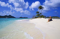 Woman sunbathing on a white sandy beach watching a catamaran, Carriacou, Caribbean.