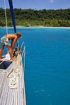 Man looking overboard at the lowering anchor aboard a cruising yacht off a sandy beach.