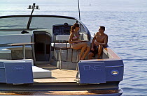 Couple sitting on bar stools on the deck of a power boat, with the transom doors open. Motorboat "One", Cantieri di Baia, off the coast of Baia, Naples, Italy.