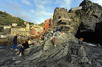 Rugged promontory with tourists resting on the water's edge in the fishing village of Vernazza, Liguria, Cinque Terre, Italy.