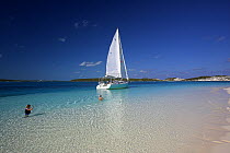 A Shannon Shoalsailor moored by a sandy beach with people in the sea. This innovative, keelless, shallow draft beachboat is designed to roam shallow waters such as these in Exuma, the Bahamas.