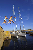 Girls jumping into water off rocks in front of two yachts at anchor.