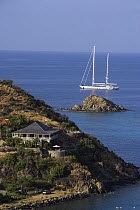 A house on the cliff past Gustavia Harbour with a yacht sailing cruising beyond, St Barthelemy, Caribbean.