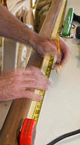 Boat-builder using a tape measure to mark out wooden boat parts with a pencil during construction of interior yacht parts.